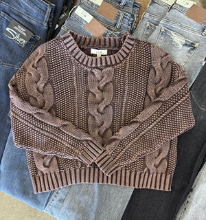 Brown cable knit sweater on a surface with jeans and other items.