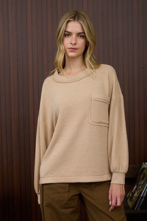 Woman wearing a beige sweater with a pocket against a wooden paneled wall.