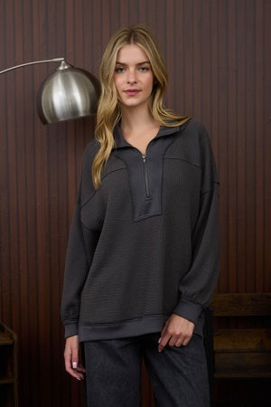 Woman wearing a dark gray hoodie and pants standing against a wooden paneled wall.
