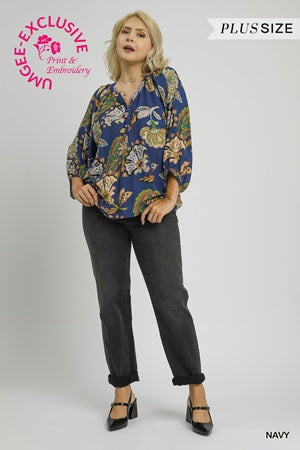 Woman wearing a blue floral blouse and black pants on a white background with branding elements.